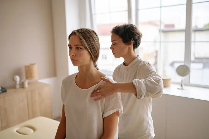 Une praticienne examine l'épaule d'une patiente debout, dans une salle lumineuse